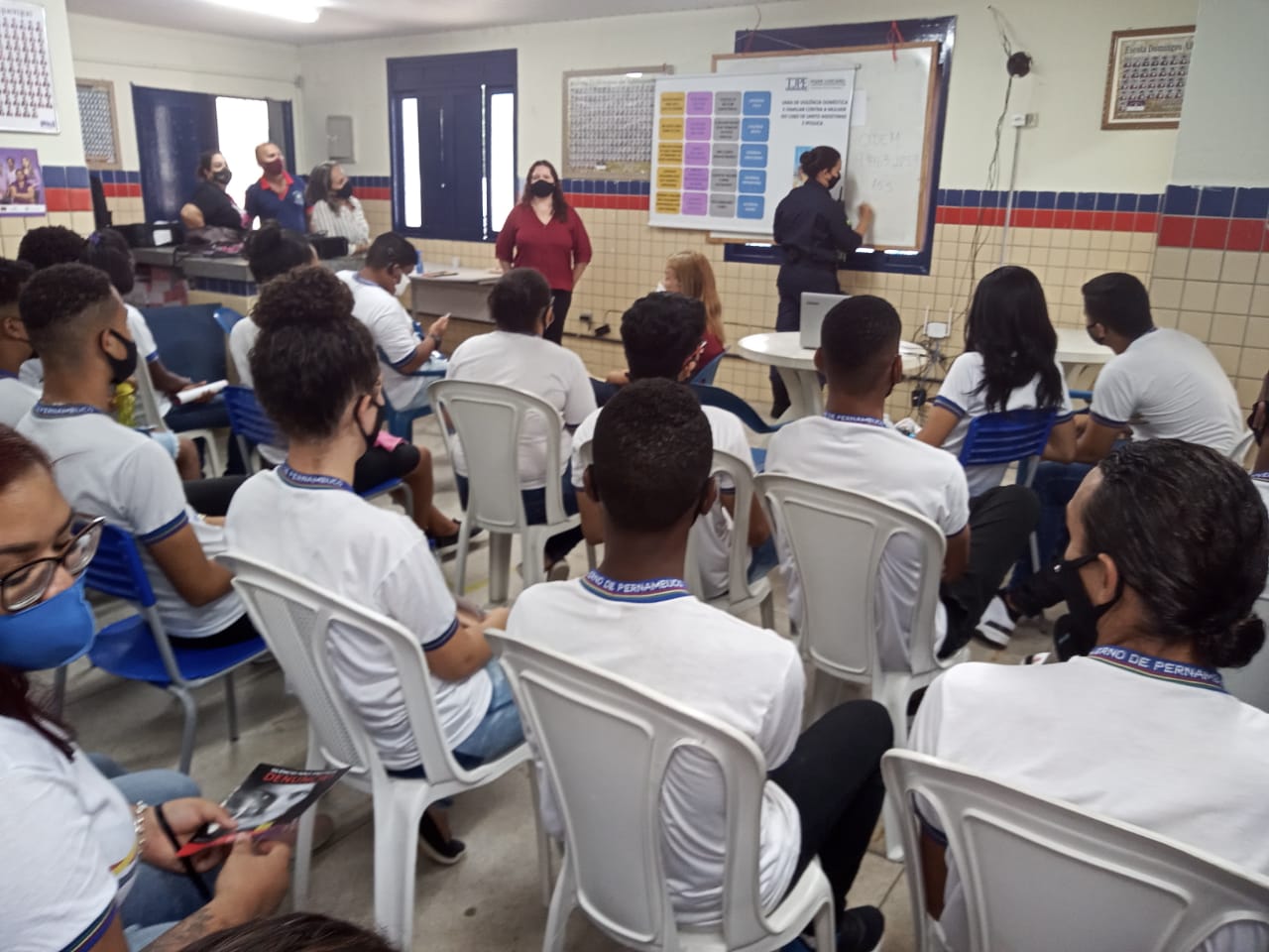 Sala de aula com um grupo de pessoas ensinando e alunos nas carteiras