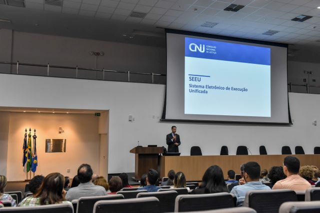 Público assiste a palestra de conferencista em um auditório com um grande telão iluminado