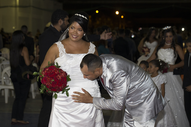 Noivo beija barriga de noiva grávida, que segura buquê de rosas vermelhas