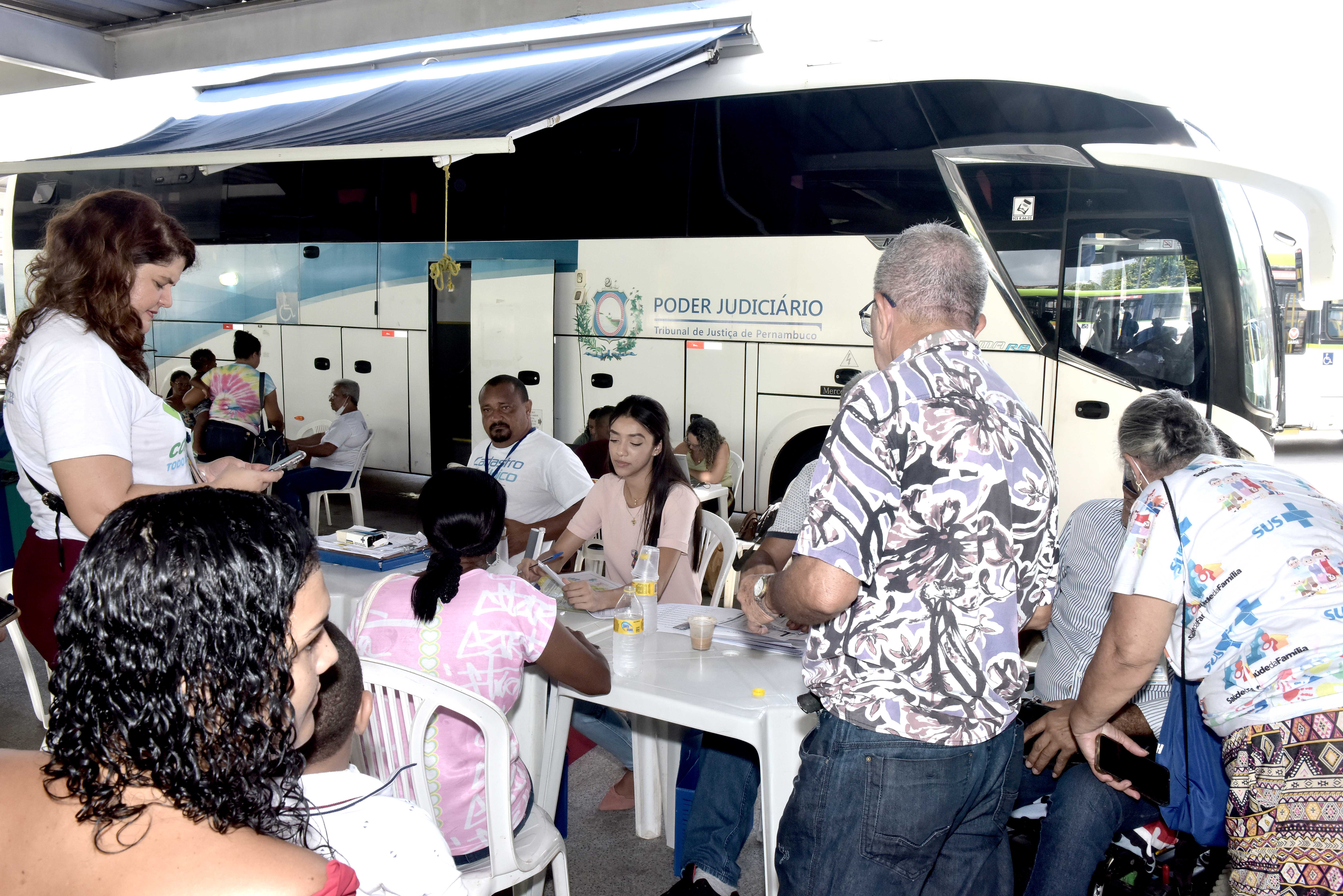 Foto mostra pessoas de pé e outras sentadas, sendo atendidas. Ao fundo aparece um ônubus na cor branca com o símbolo e o nome Poder Judiciário.