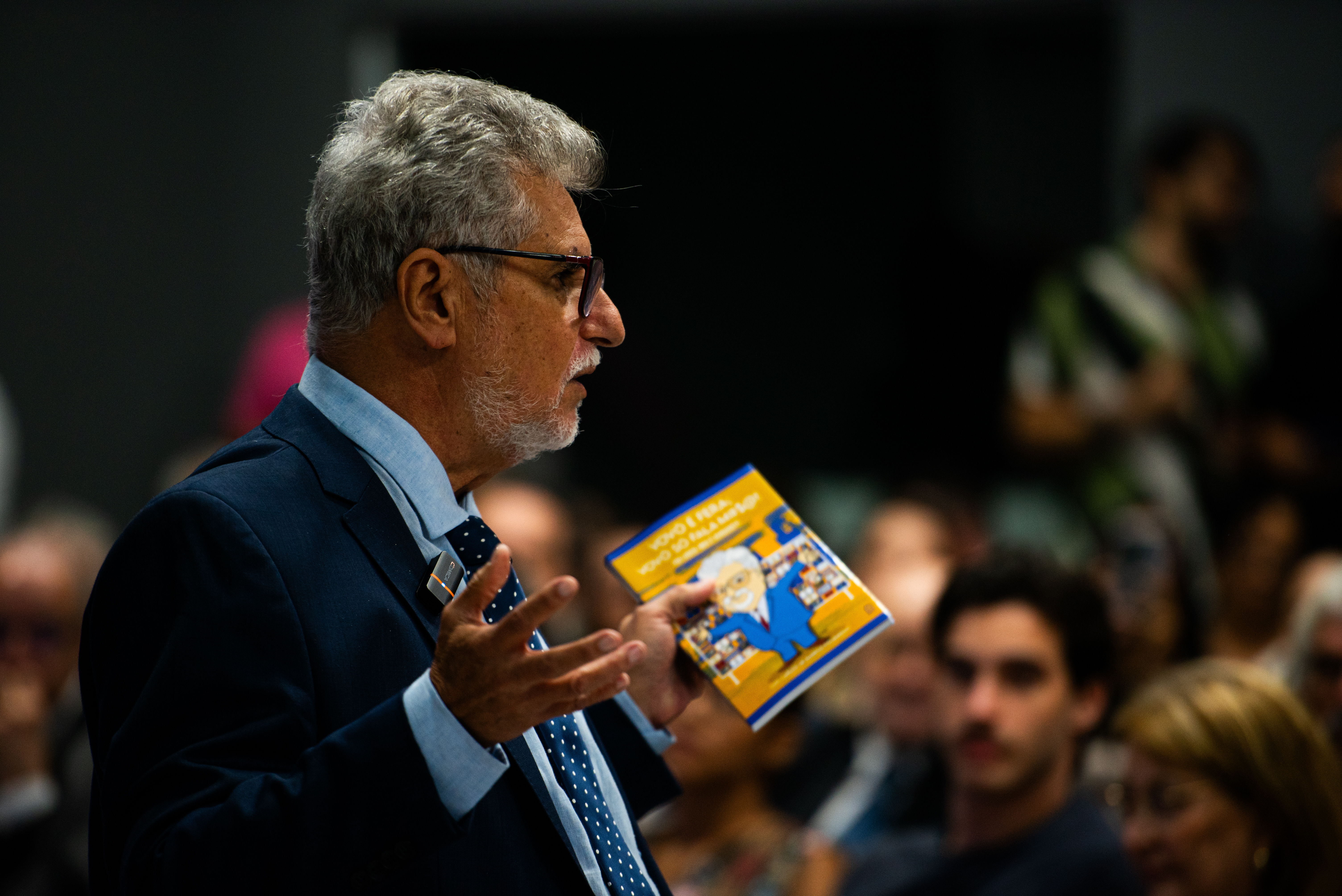 Homem em pé, gesticulando com um livro na mão, para uma plateia