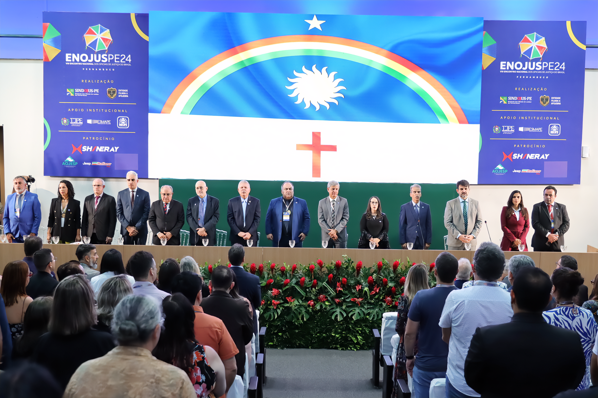 14 pessoas em pé atrás de uma mesa com a bandeira de Pernambuco cobrindo toda a parede atrás deles