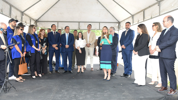 Pessoas lado a lado, em pé sobre palco, observando uma mulher discursando.