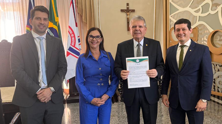 À frente das bandeiras de Pernambuco, do Brasil e do TJPE, três homens e uma mulher estão lado a lado e sorridentes. Vestem roupas sociais. Ao centro, o presidente do TJPE segura um convite.