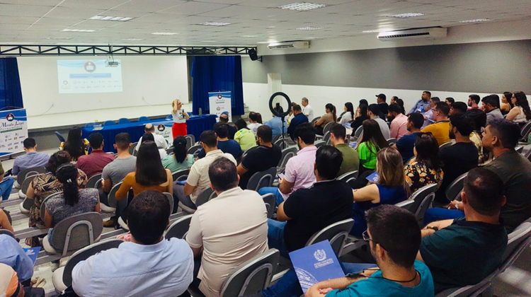 Em auditório, pessoas sentadas assistem à apresentação, em telão; e fala da coordenadora adjunta do Moradia Legal, Sara Lima, presencialmente.