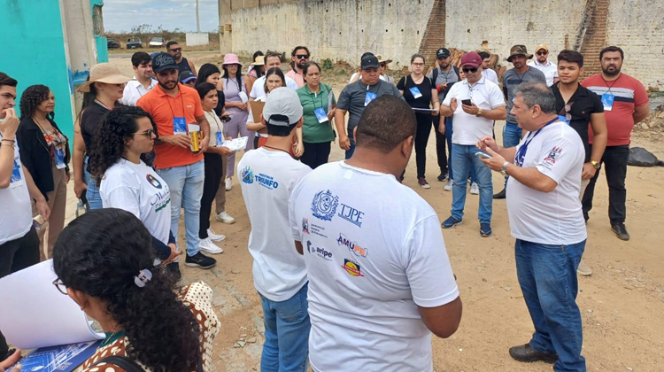 Diversas pessoas em pé, em rua de barro, perto de casas, prestam atenção a orientações passadas pelo professor Silvio Garnés, para início das atividades de regularização de imóveis.