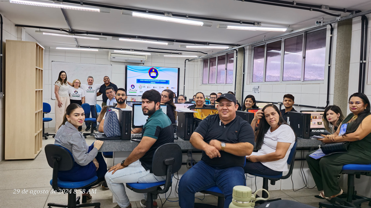 Pessoas sentadas e em pé assistem à aula em laboratório de informática.