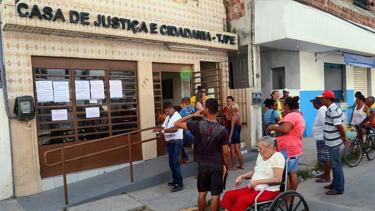 Foto de uma casa escrito acima em letras grandes e pretas "Casa de Justiça e Cidadania - TJPE", com azulejos na cor branca e marrom, portões marrom e com rampa de acesso. Na frente da casa, aparecem várias pessoas da comunidade e uma senhora cadeirante.