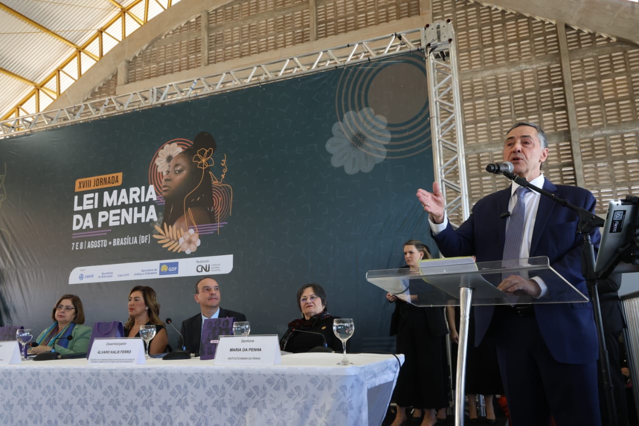 O presidente do STF e do CNJ, Ministro Luís Barroso, fala em pé no evento XVIII Jornada Lei Maria da Penha. Ele está em pé, enquanto outros participantes estão sentados.